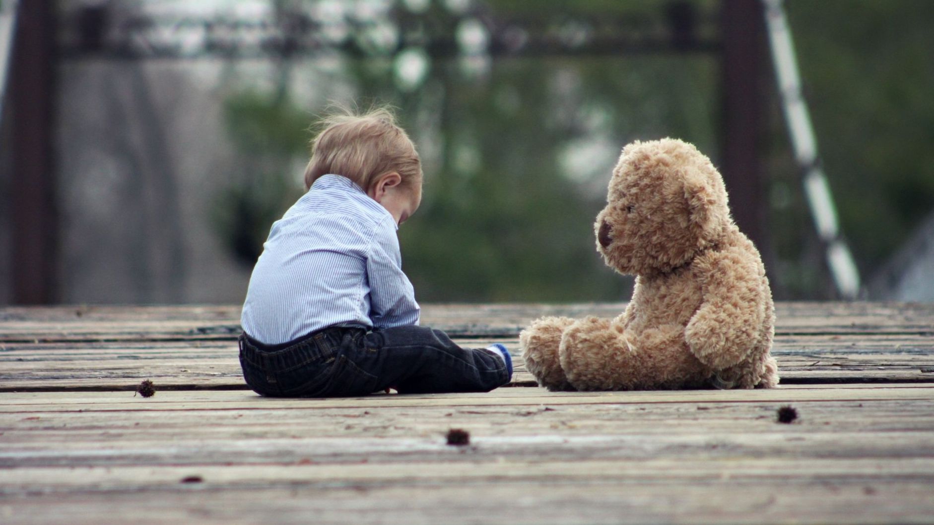 a little boy is sitting next to a teddy bear on a wooden deck .