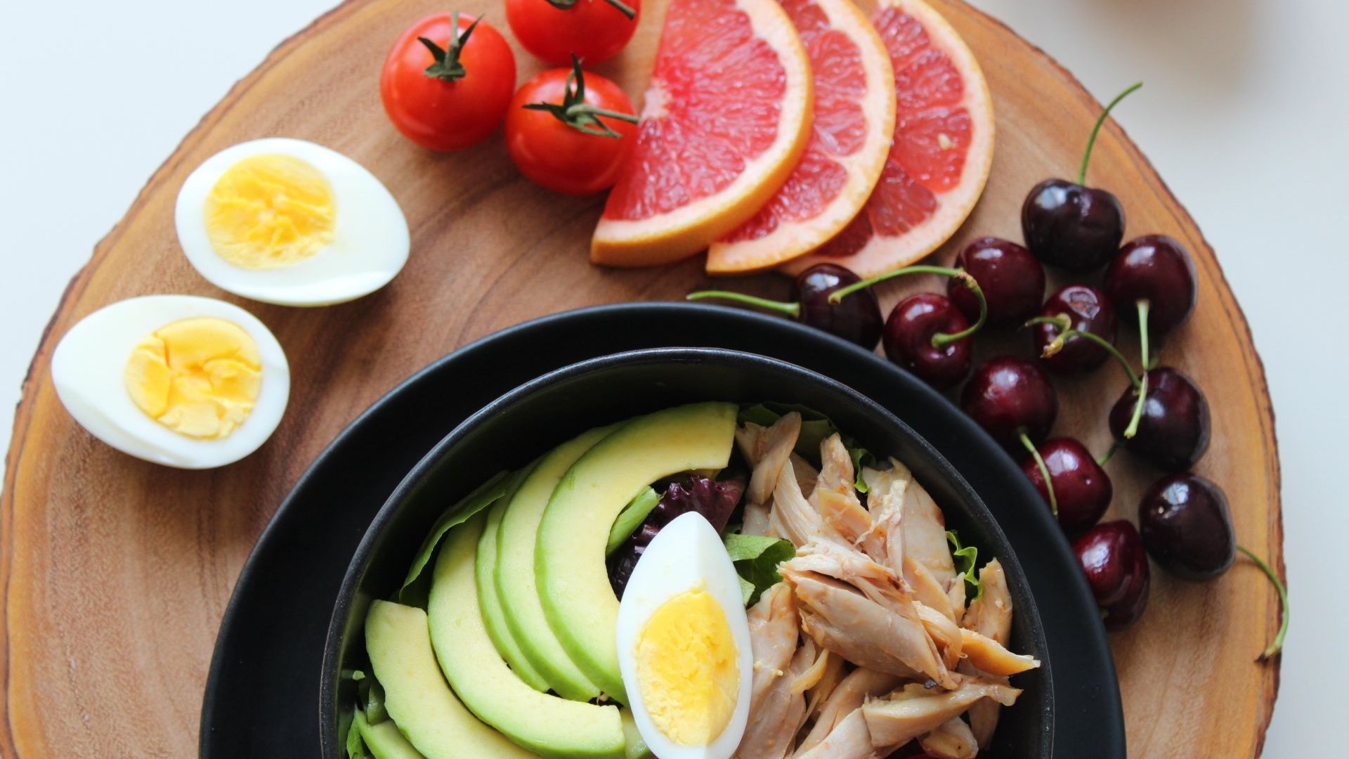 a bowl of food with avocado , eggs , grapefruit and cherries on a wooden cutting board .