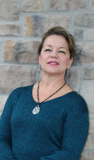 A woman in a blue sweater and necklace is standing in front of a brick wall.