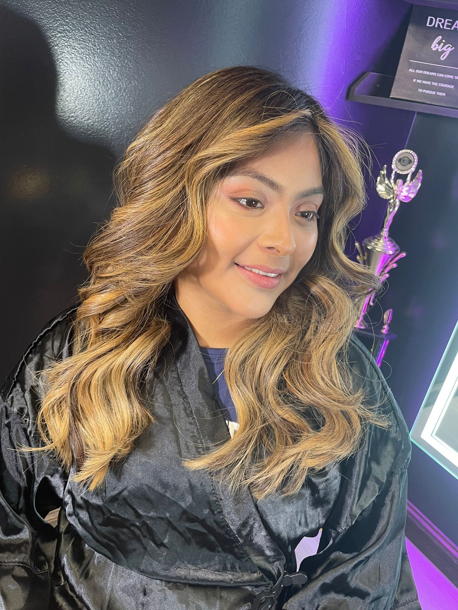 Woman with wavy, highlighted hair smiling, wearing a black robe in a salon setting.