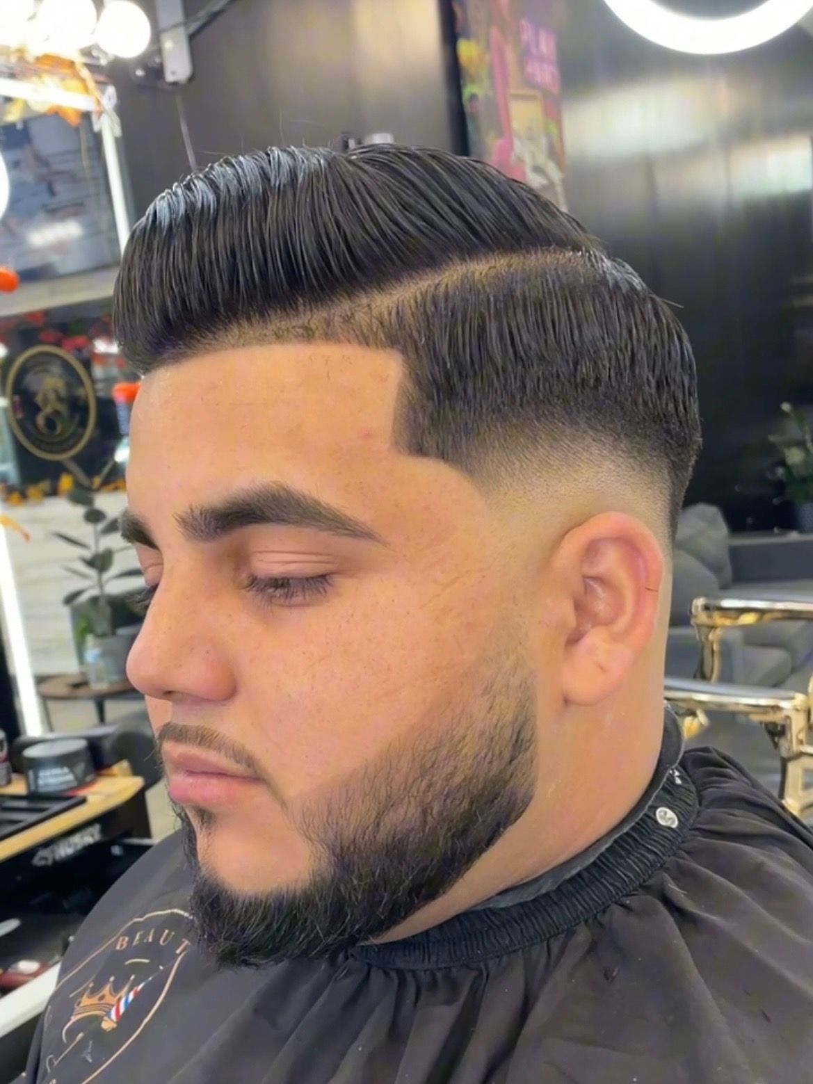 Man with a faded haircut, styled pompadour, and trimmed beard. He’s wearing a barber's cape, indoors.
