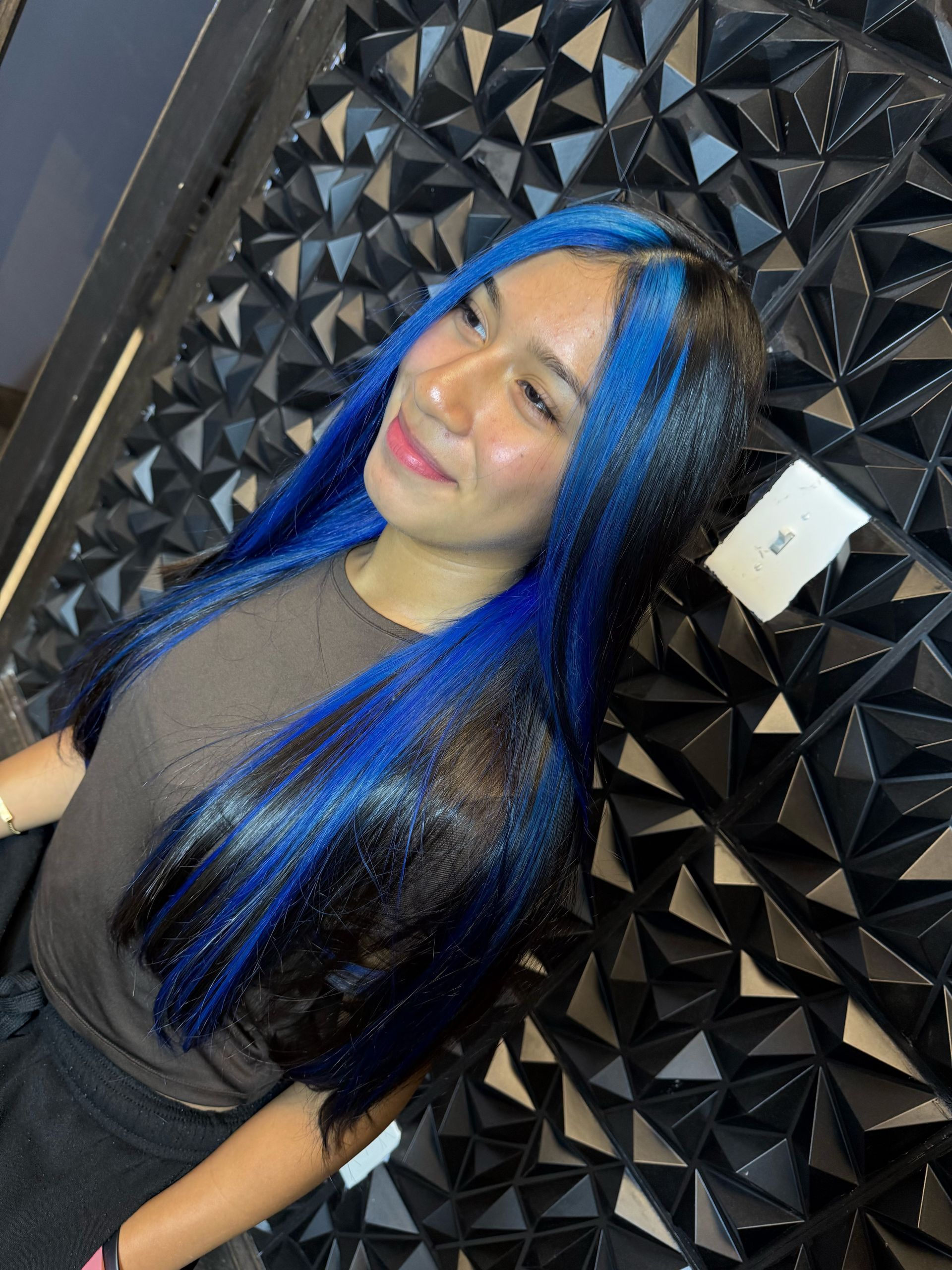 Woman with black hair and bright blue highlights, smiling, against a textured black wall.