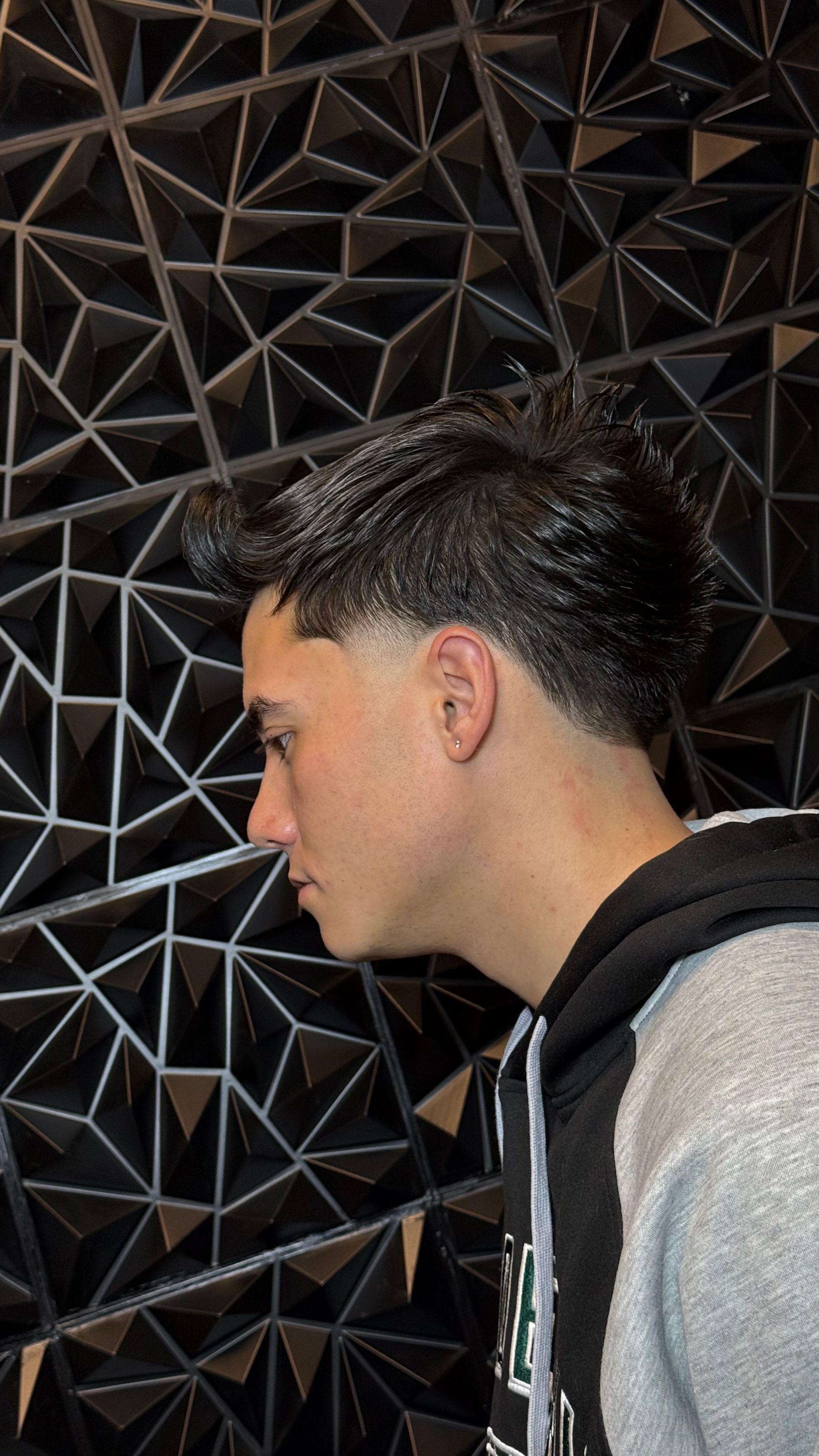 Person with a fade haircut and dark hair styled back, wearing a hoodie, posing against a geometric black wall.