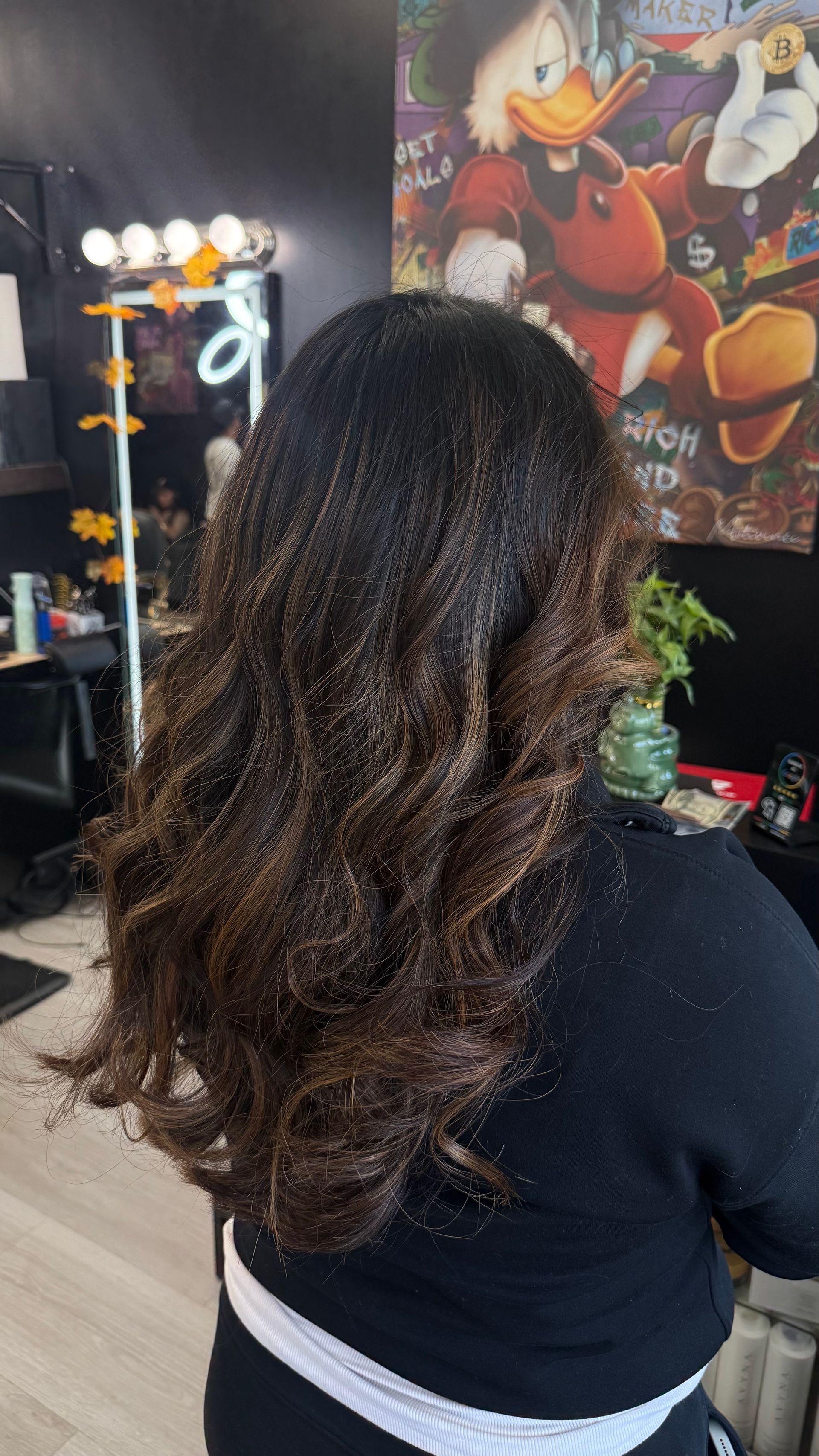 Woman with dark brown hair, styled in loose curls. Hair has light brown highlights. The background is a hair salon.