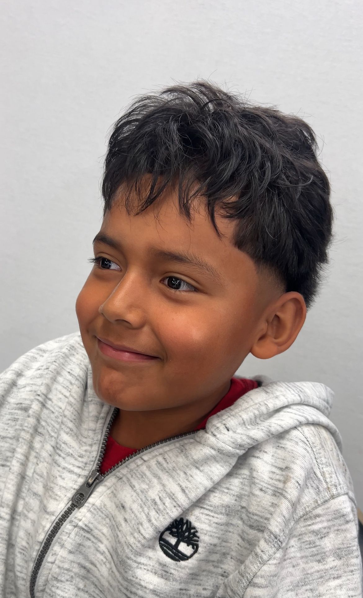 Boy smiling, wearing gray zip-up hoodie, dark hair with fade haircut.