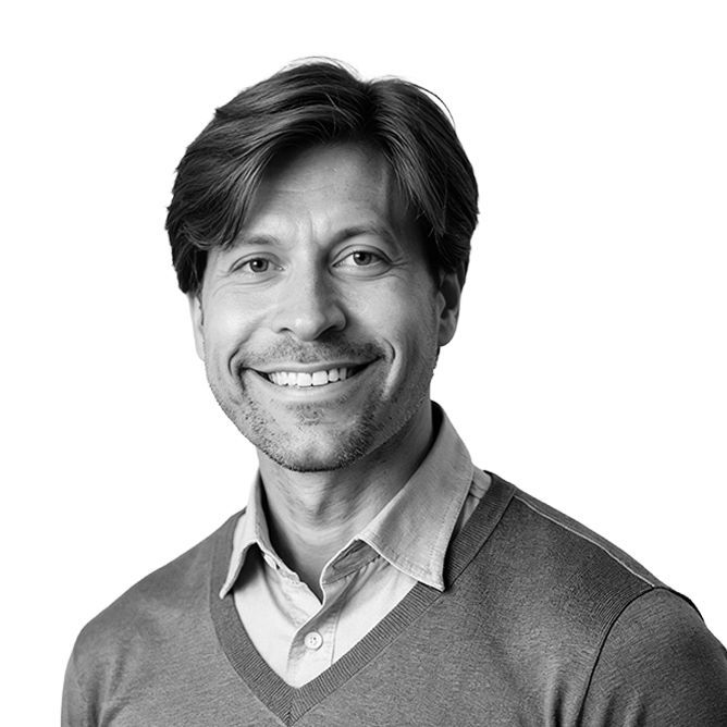 Man with short hair smiles, wearing a light-colored shirt under a gray sweater.