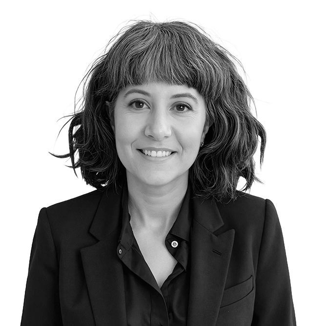 Woman with wavy, dark hair and bangs smiles, wearing a black blazer and collared shirt.