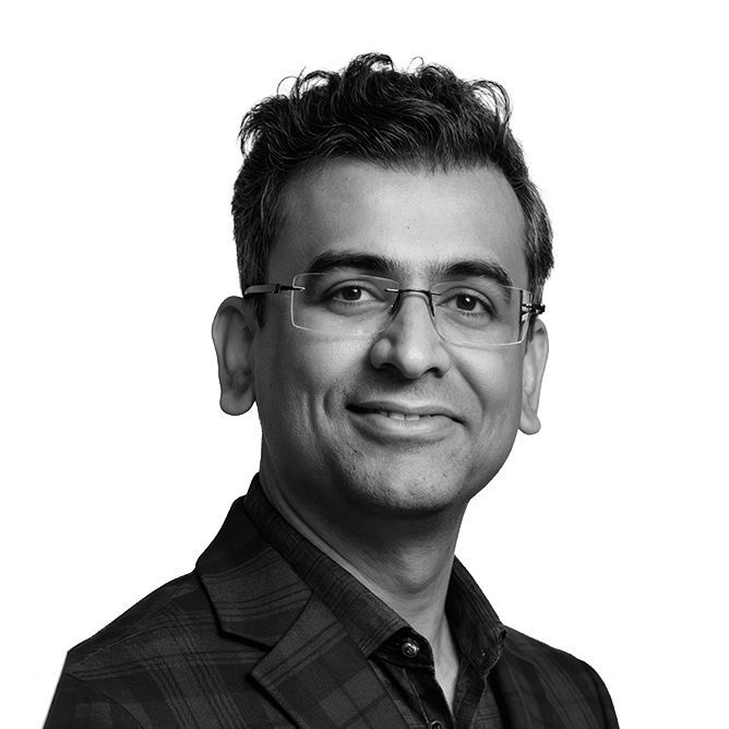 Man wearing glasses smiles, in a blazer, indoors. Black and white headshot.