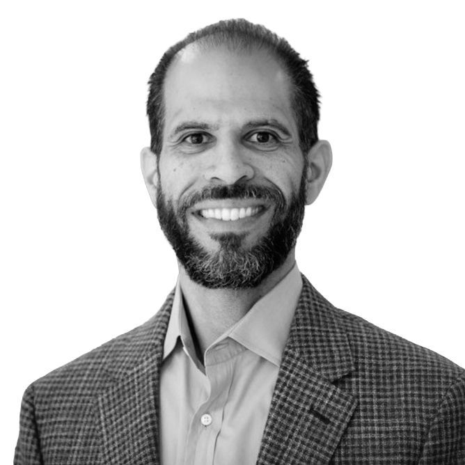 Man with a beard smiling, wearing a blazer and button-down shirt, headshot in grayscale.