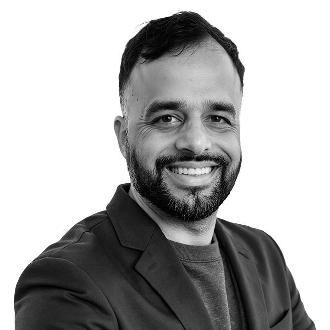Man with a beard smiling, wearing a dark blazer over a gray top, in a studio setting.