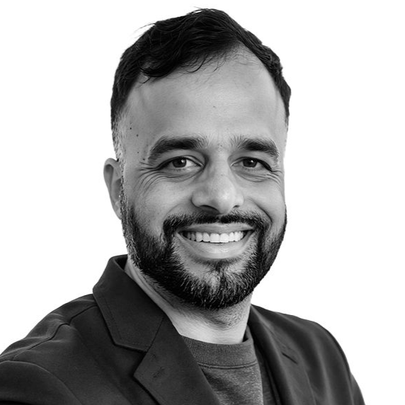 Man with a beard smiling, wearing a dark blazer over a gray top, in a studio setting.
