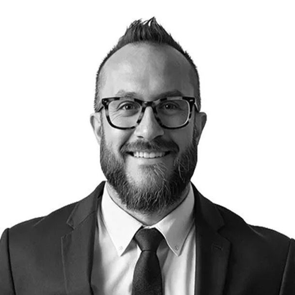 Man in suit and glasses smiles, with dark beard and hair, white background.