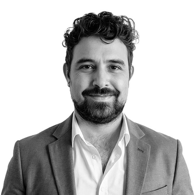 Man with curly hair and beard, wearing a blazer over a white shirt, smiling.