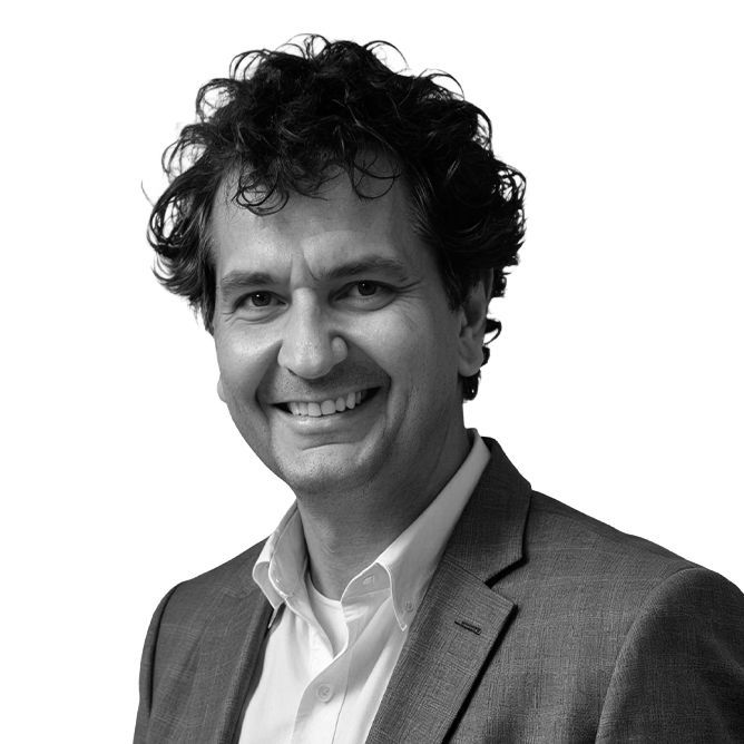 Man with curly dark hair smiles, wearing a blazer and white shirt, isolated on white background.