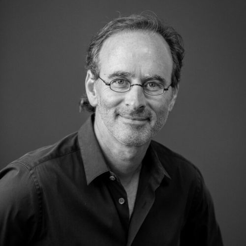 Man with glasses smiles, looking at camera. Dark shirt, monochrome studio portrait.
