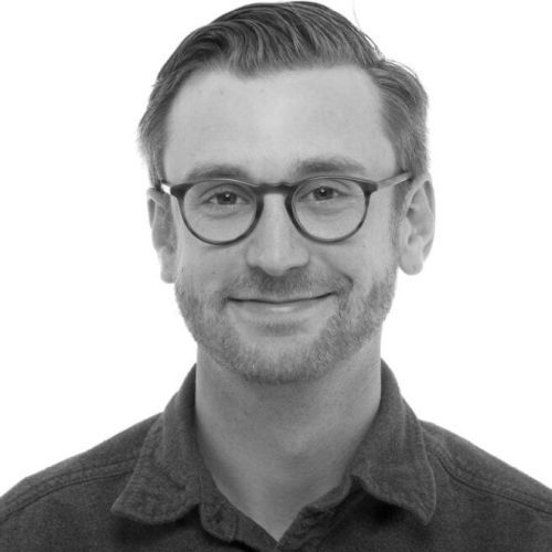 Man with glasses smiles, looking forward; wearing a dark button-down shirt.