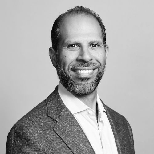 Man in a blazer smiling at the camera, short dark hair, full beard, neutral background.