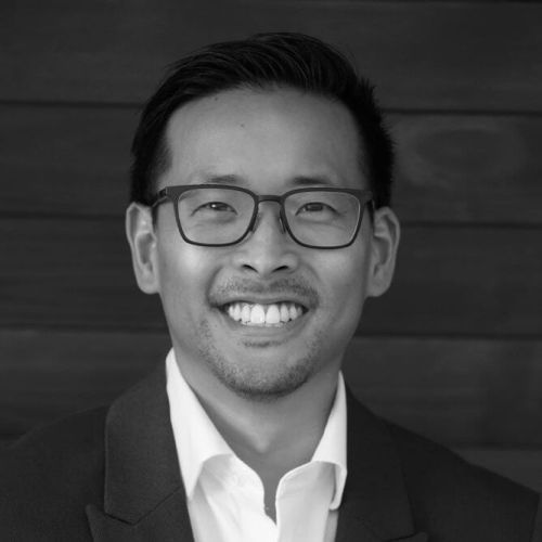 Smiling man with glasses and blazer against a wood background.