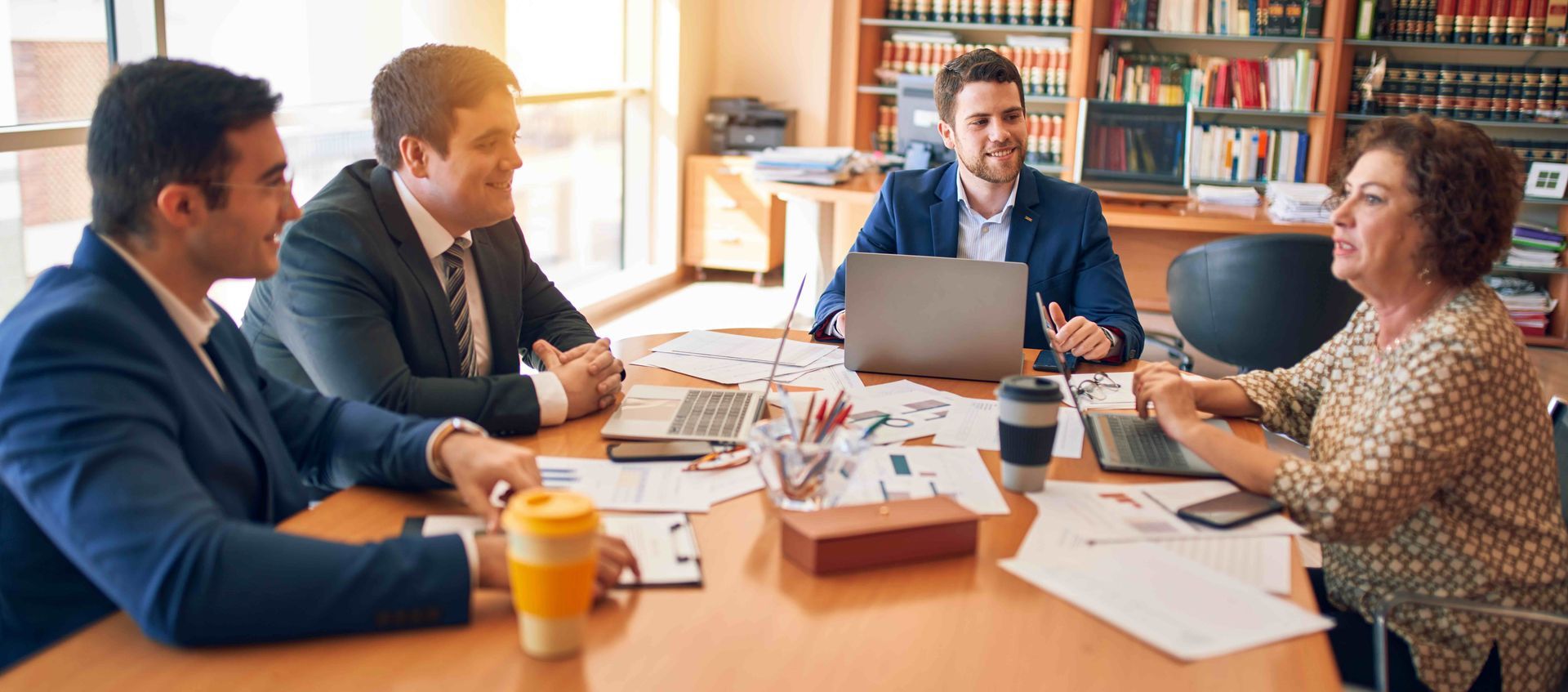 Four people in business attire seated around a table, discussing documents. Sunlight streams in from a window, illuminating the room.