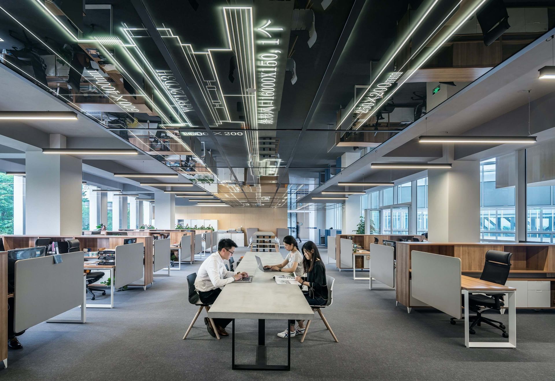 A bright, modern open-plan office with rows of desks, gray carpeting, and exposed ceiling lighting.