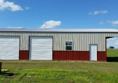 A large metal building with two garage doors and a window is sitting in the middle of a grassy field.