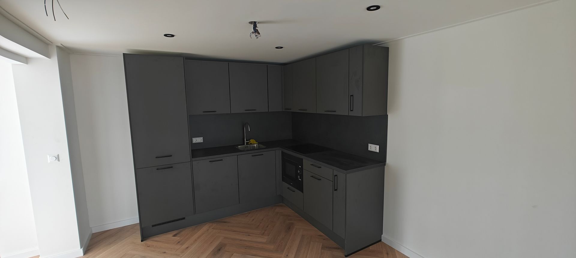 Een moderne, L-vormige keuken met mat donkergrijze kasten, donkere werkbladen en een lichte visgraatparketvloer.