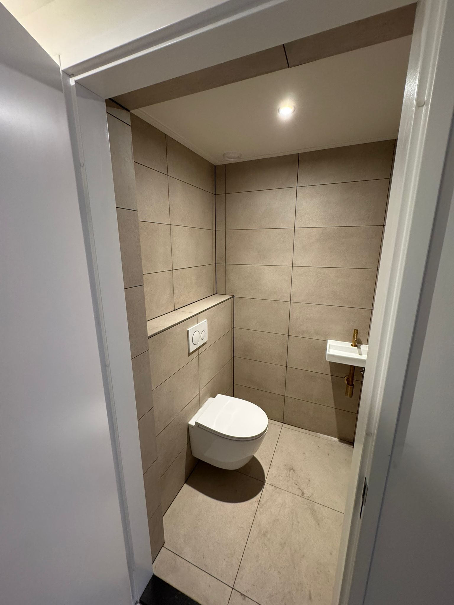 Een kleine, moderne toiletruimte met beige betegelde wanden en vloer, voorzien van een wandtoilet en een kleine hoekwastafel.