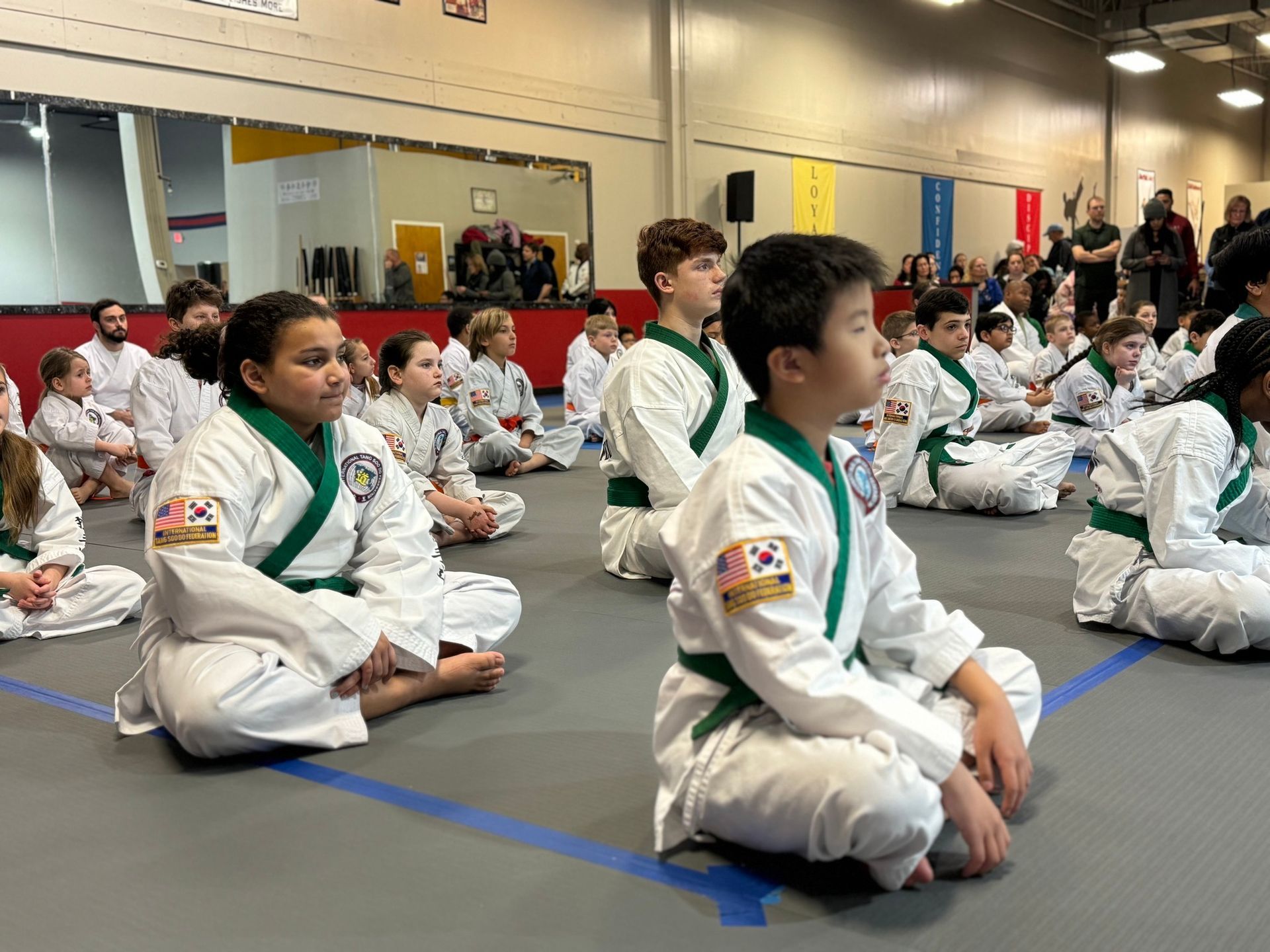 A group of children in karate uniforms are sitting on the floor in a gym.