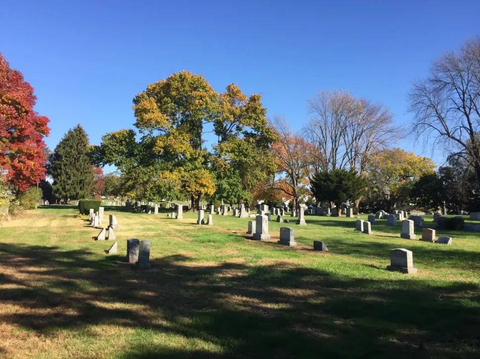 Cemetery with many headstones on a grassy hill, surrounded by trees with colorful fall foliage under a clear blue sky.