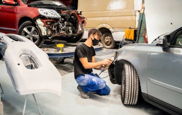 A man works at a car collision repair shop, surrounded by tools and car parts, focused on repairs.