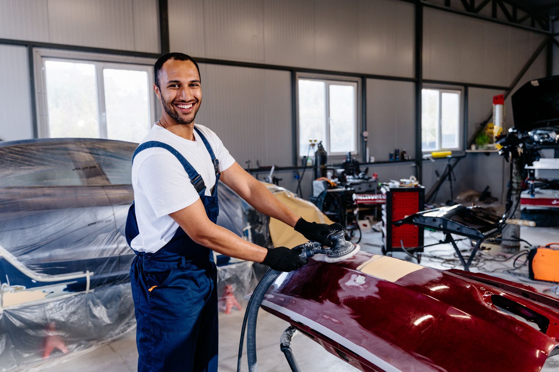 Smiling mechanic polishing red car bumper inside professional car collision repair shop.