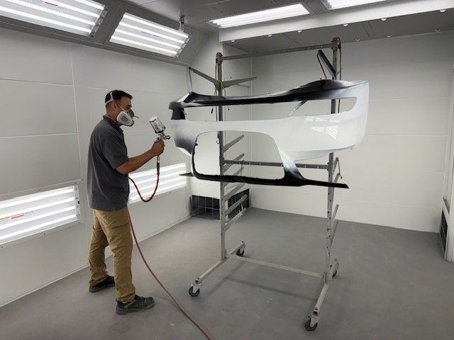 A man is spray painting a car bumper in a paint booth.