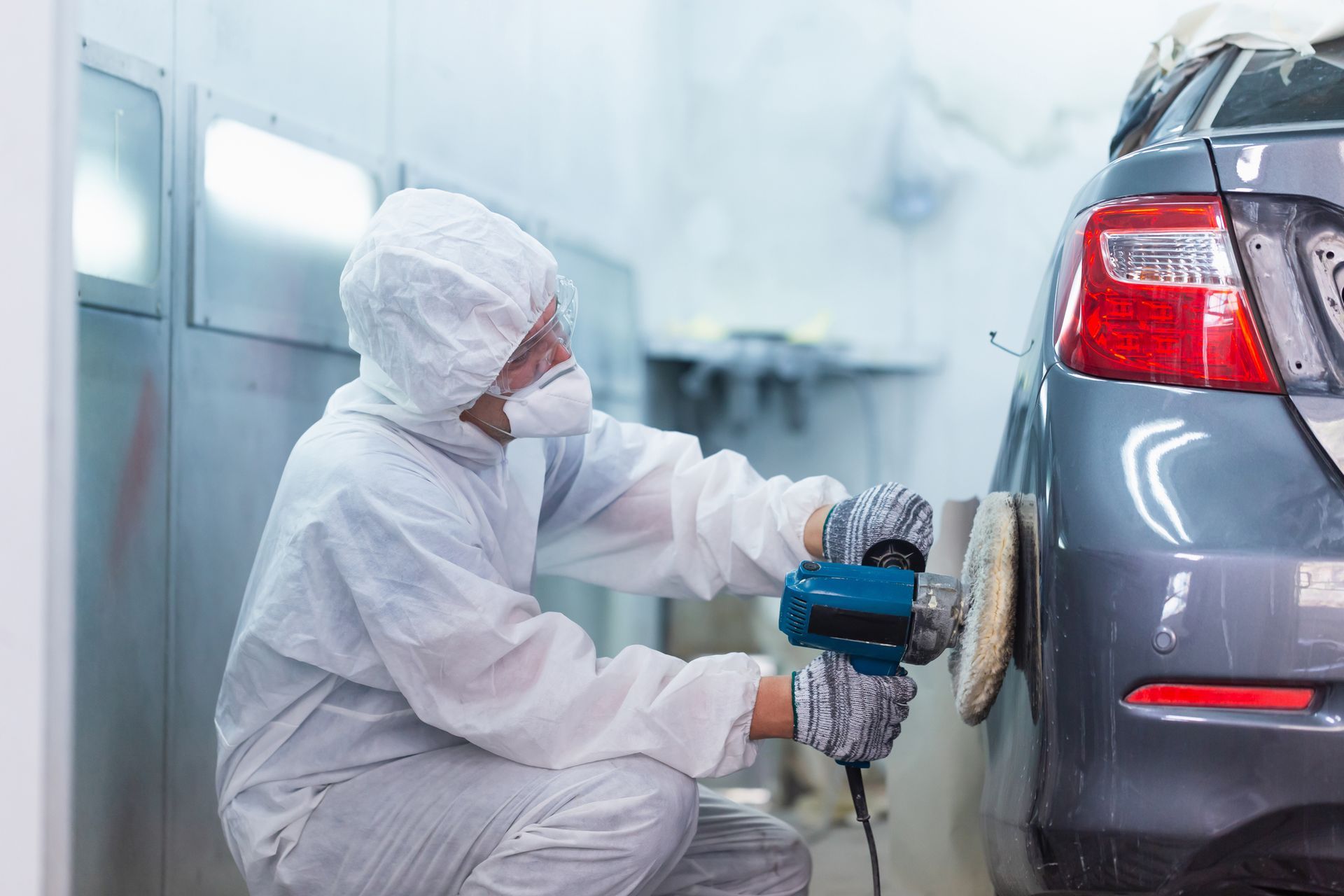 Technician in protective suit polishing rear bumper at professional car collision repair shop.