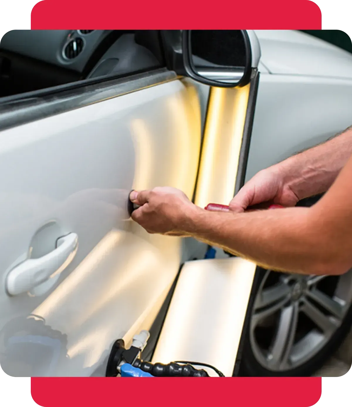 Person using tools to remove a dent from a white car door, illuminated by a light.