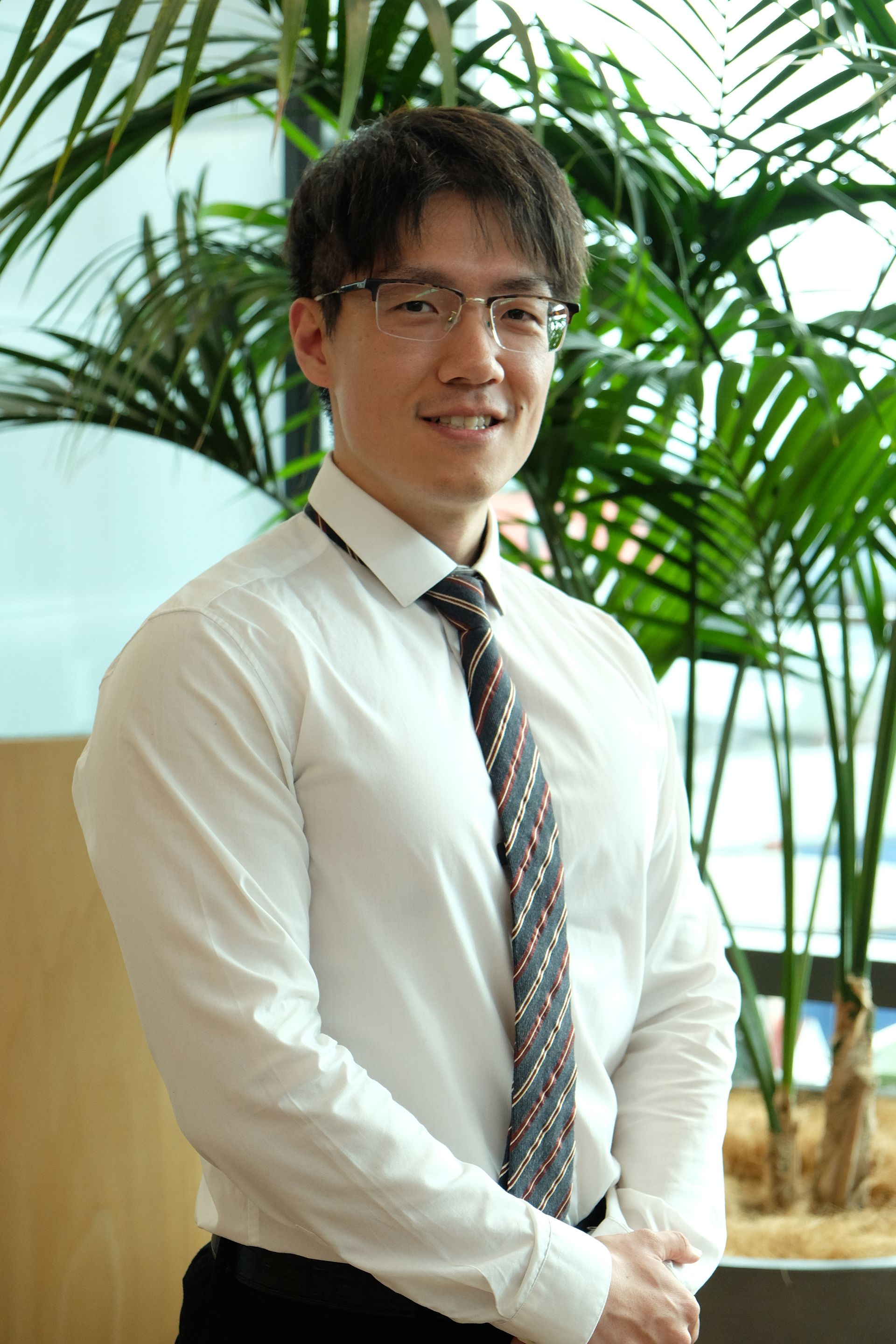 Dr Anbin Li  in white shirt and patterned tie standing indoors near green plants, smiling gently.