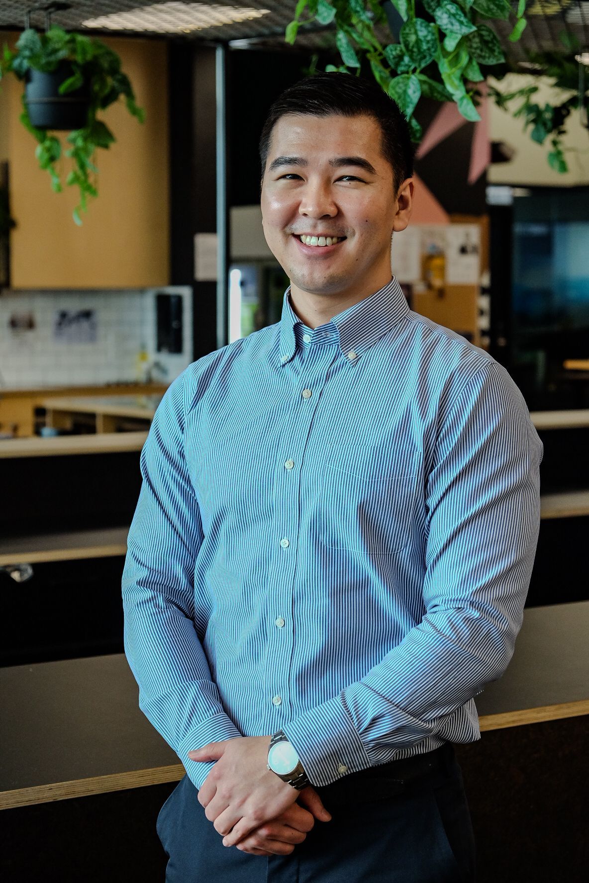 Andrew Chen in a striped shirt standing indoors near a counter and hanging plants.