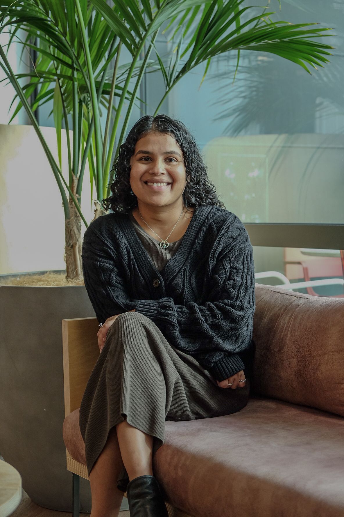 Bidara Pathirage- Principal Engineer seated on a brown couch beside a tall potted palm in a bright room.