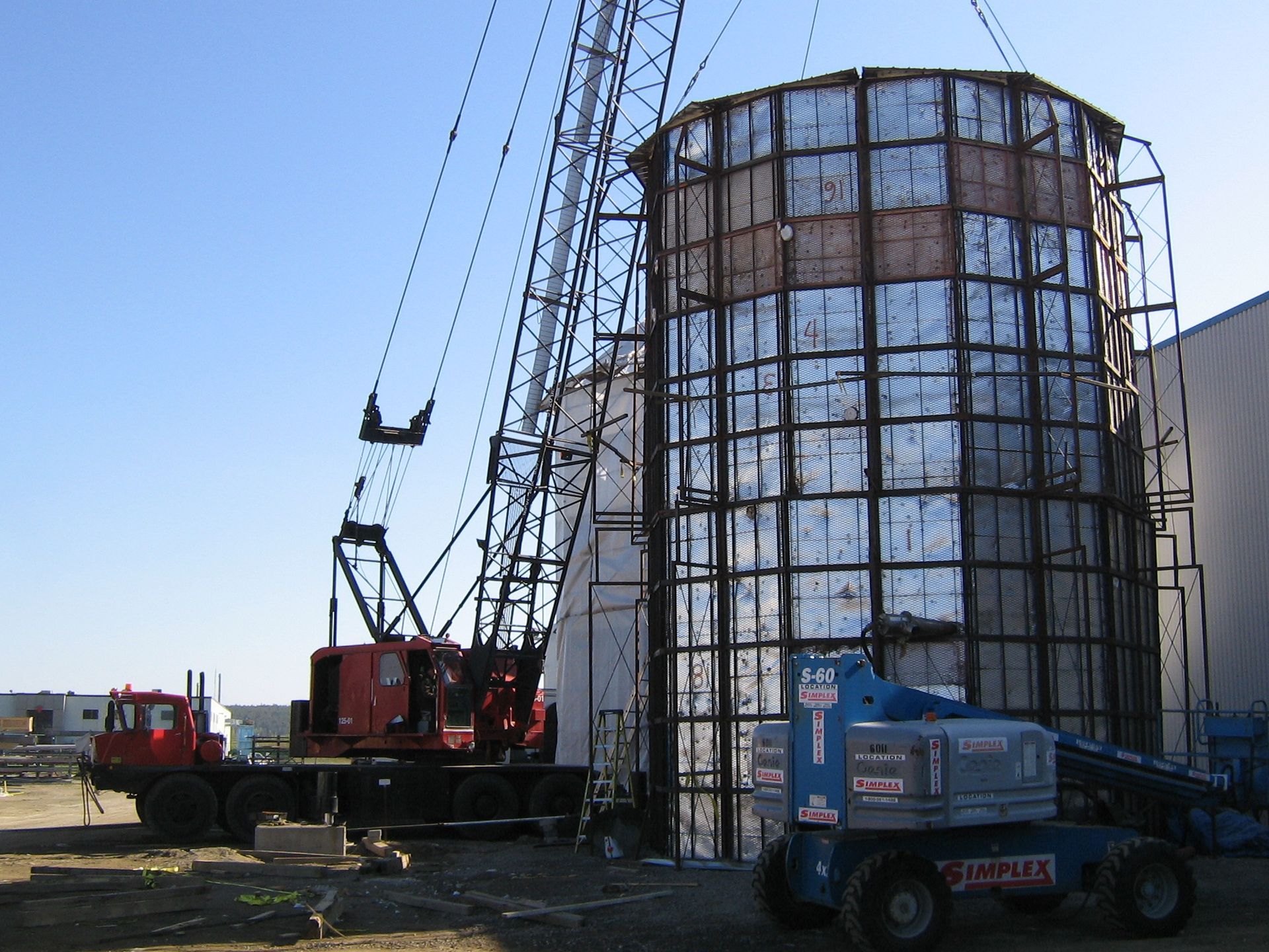 Grue soulevant une grande structure métallique ; chantier de construction. Grue rouge, ascenseur bleu ; journée ensoleillée.