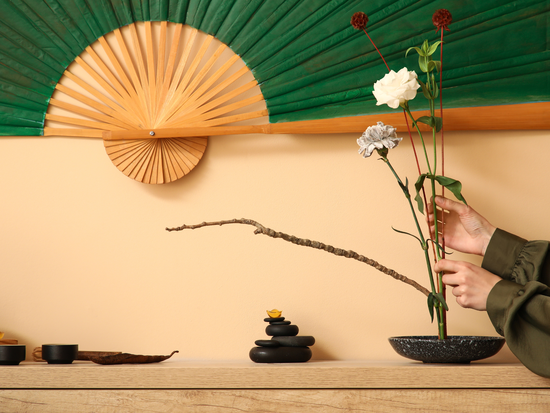 Person arranging flowers in a black vase on a shelf, green and beige fan in background.