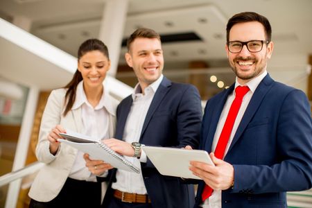 Three business professionals smiling in an office, holding a clipboard and tablet.
