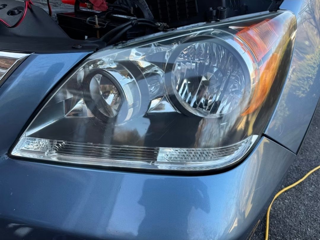 Close-up of a car's headlight, showing clear lenses and a partially open hood. The car is blue.