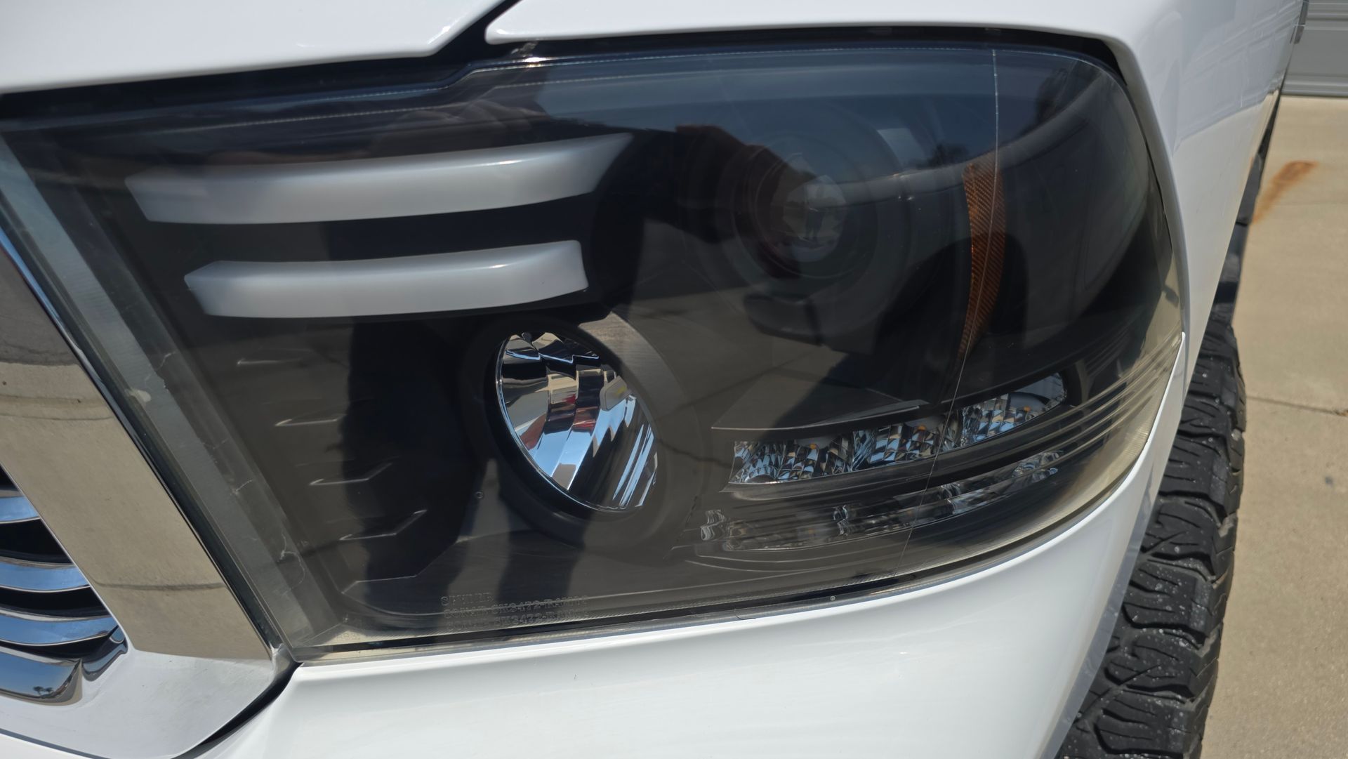 Black tinted headlight on a white truck; close-up shot showing a light, grill, and tire.
