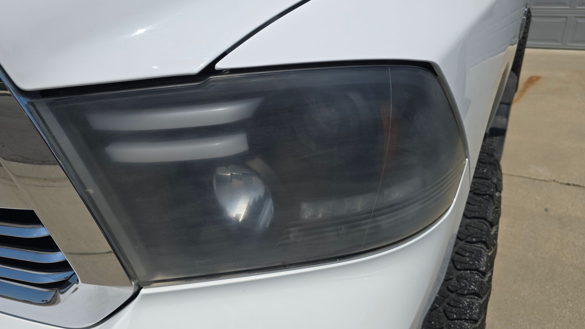 Close-up of a white truck's front, showing a blacked-out headlight. The headlight is slightly foggy.
