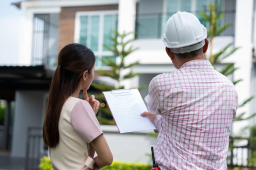 Two people reviewing a clipboard outside a house, one wearing a hard hat, with a car and greenery in the background
