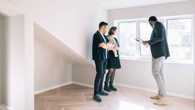 A real estate agent talks to a couple holding a baby while showing them an empty, modern room with slanted ceilings.