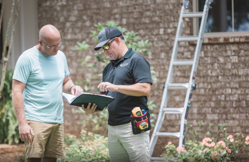 Two men reviewing plans outdoors beside a ladder and stone wall