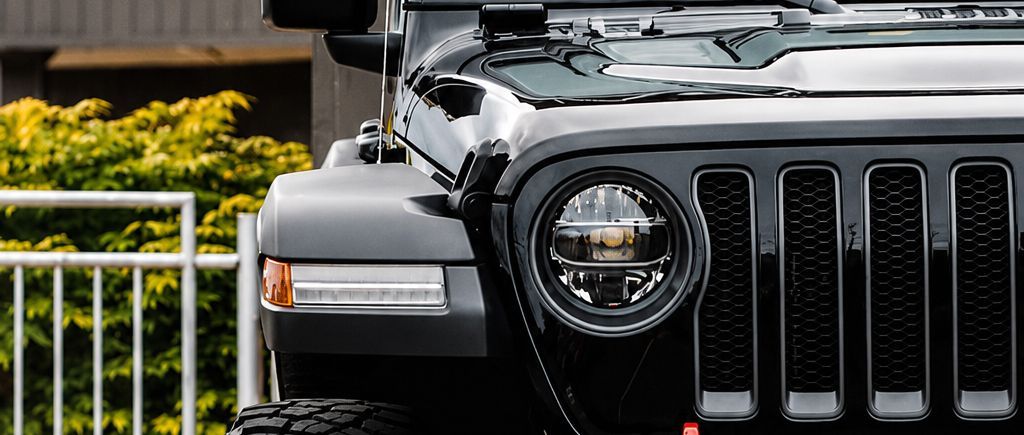 Close-up of a black Jeep front grille and headlight beside a white fence and greenery