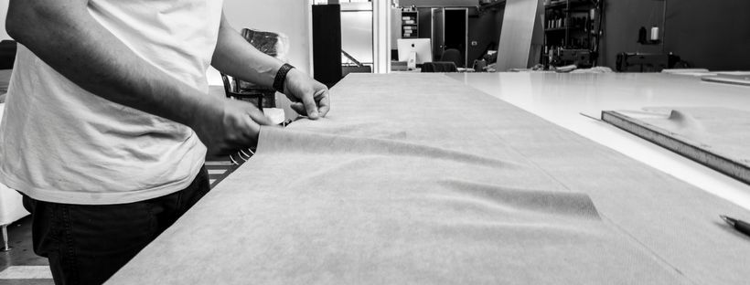 Persona che usa le forbici per tagliare la carta su un grande tavolo, con un righello e una matita visibili sullo sfondo.