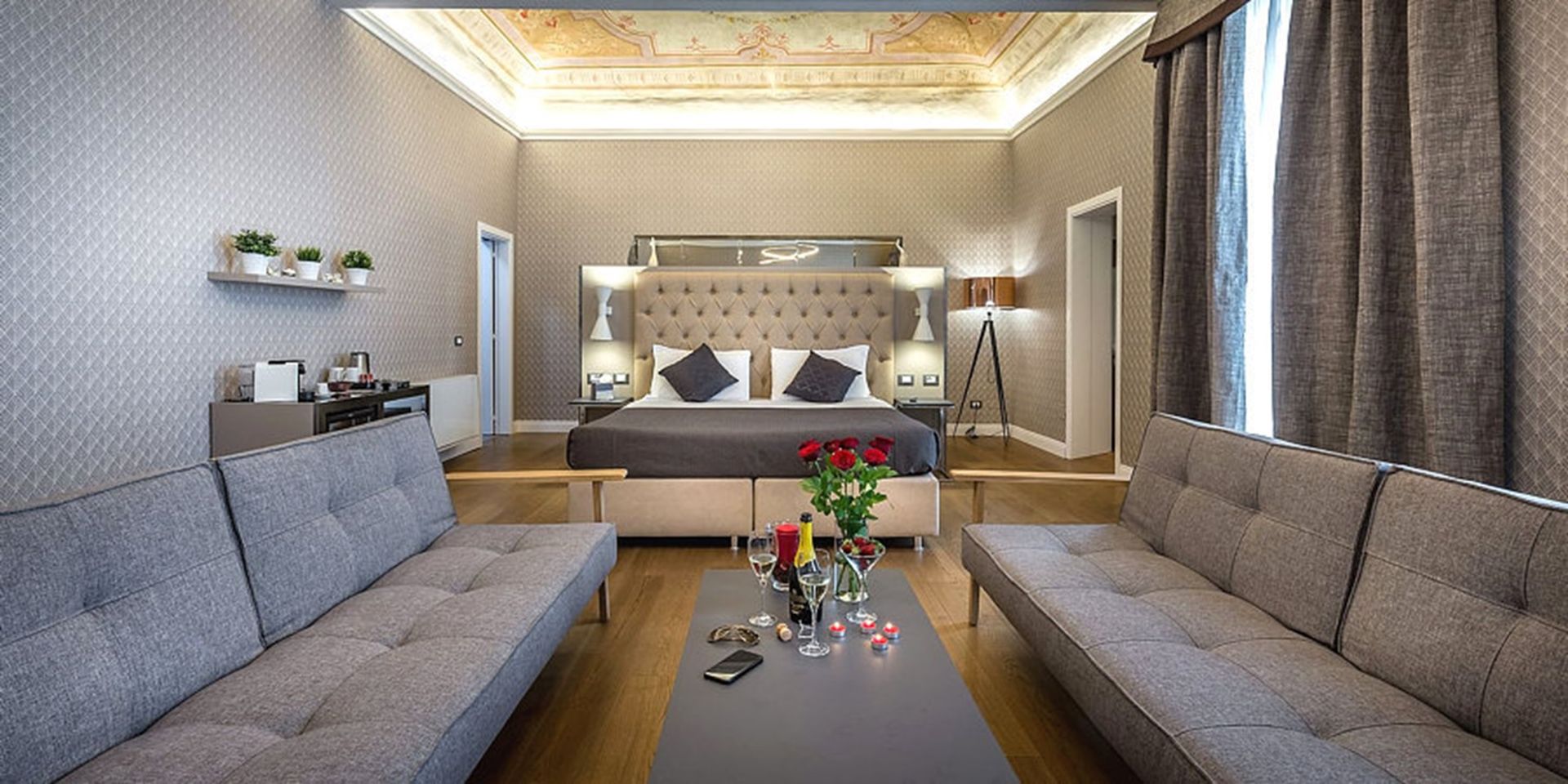 Una suite di lusso con un letto, due divani e un tavolino con fiori e bevande. Le pareti sono decorate con un motivo testurizzato.