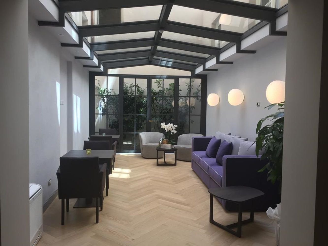 Salotto soleggiato con tetto in vetro, pareti grigie, divano viola, poltrone, tavoli scuri e piante. Ampie porte a vetri conducono a uno spazio verde.